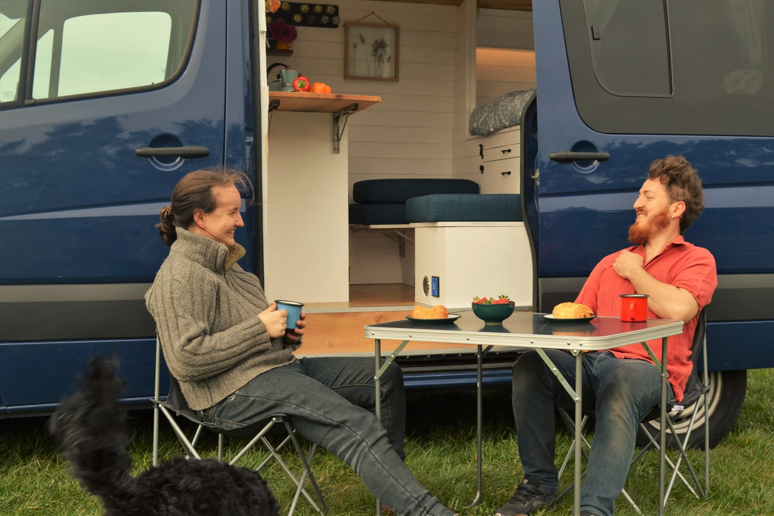 Two people with tea beside an open campervan on the grass.